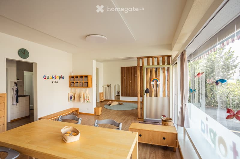 Bright, open room with wooden flooring, large window with view, and various furniture including a table, chairs, bench, and storage unit.