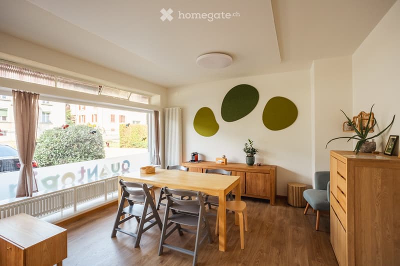 Bright, modern room with wooden flooring, large window with view, round wall decorations, and a mix of wooden and grey furniture including a dining table, chairs, and a cabinet.