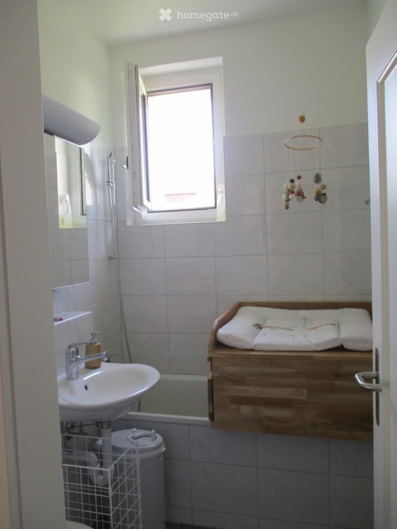 Bathroom with white tiled walls, a sink with a faucet, a mirror, a toilet, a bathtub, a wooden shelf with a cushioned surface, and a hanging toy mobile.