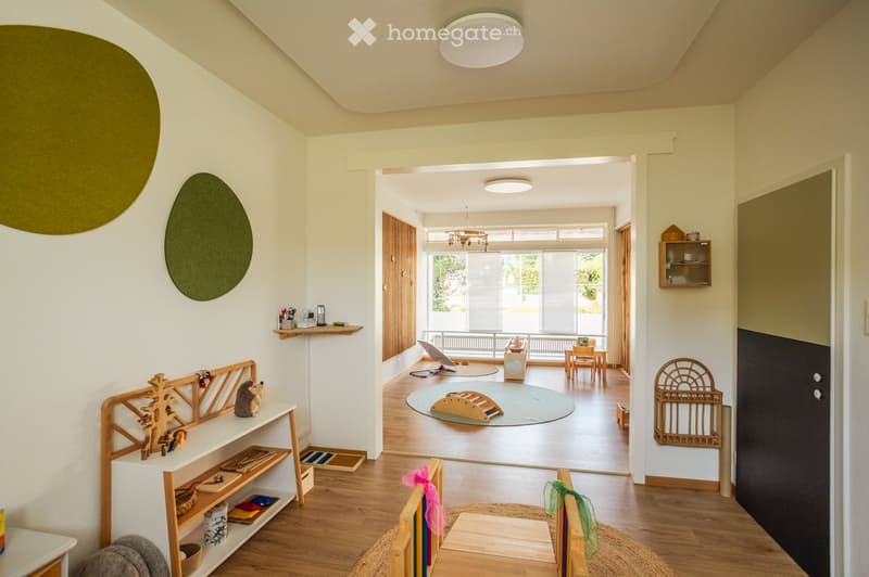 Bright, open-plan room with wooden flooring, large windows, and modern decor. Features include a shelf with toys and books, a small table with chairs, a round rug, and a cabinet with glass doors.