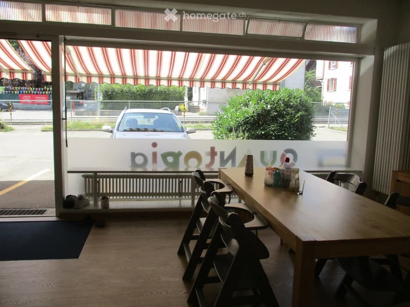 A small, well-lit room with a wooden table and chairs, a few bottles and cups on the table, and a view of the street through large windows.