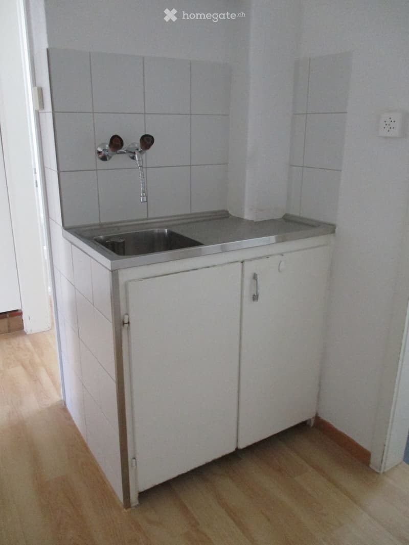 A small kitchen with a white tiled backsplash, a stainless steel sink, and white cabinets with silver handles. The floor is wooden.