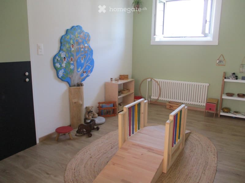 A well-lit children's room with wooden flooring, a colorful tree mural, a small wooden bed, a stool, a teddy bear, and various toys and shelves.