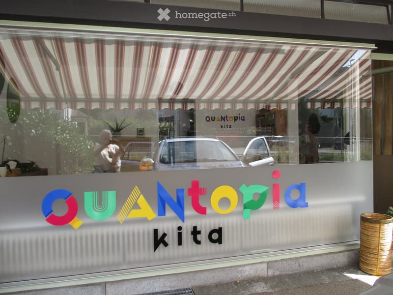 Storefront with large glass windows and colorful signage reading "QUANTOPiA kita". Visible interior includes a potted plant, a person taking a photo, and a car reflected in the glass.