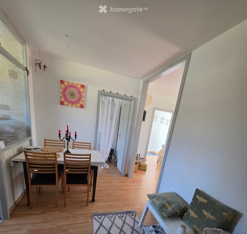 Dining area with a wooden dining table, chairs, a candle holder, a bench with pillows, and a rug on the floor. The room has light-colored walls, wooden flooring, and large windows with blinds.