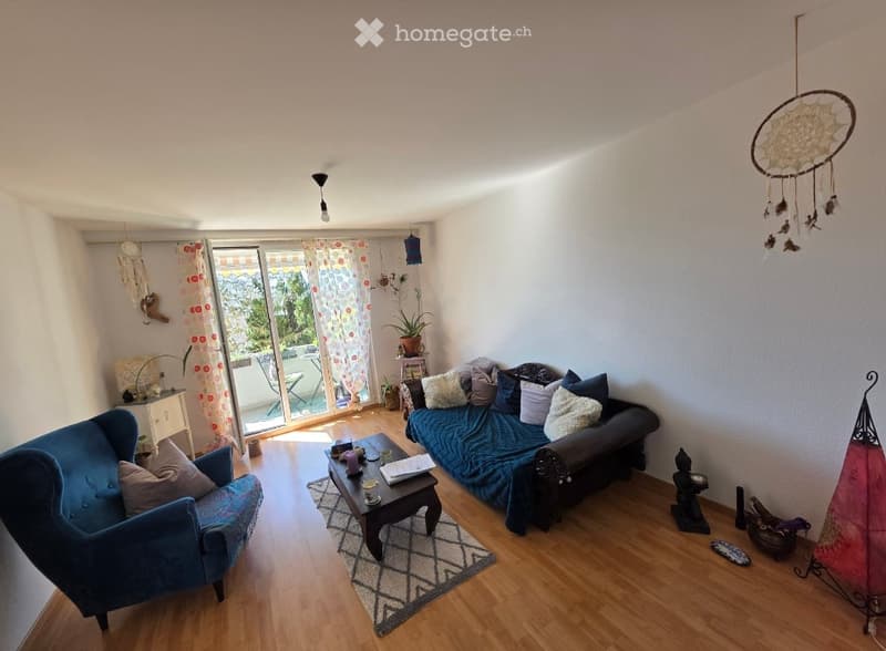 Living room with wooden flooring, large window with balcony access, sofa, armchair, coffee table, and decorative items including a dreamcatcher and plants.