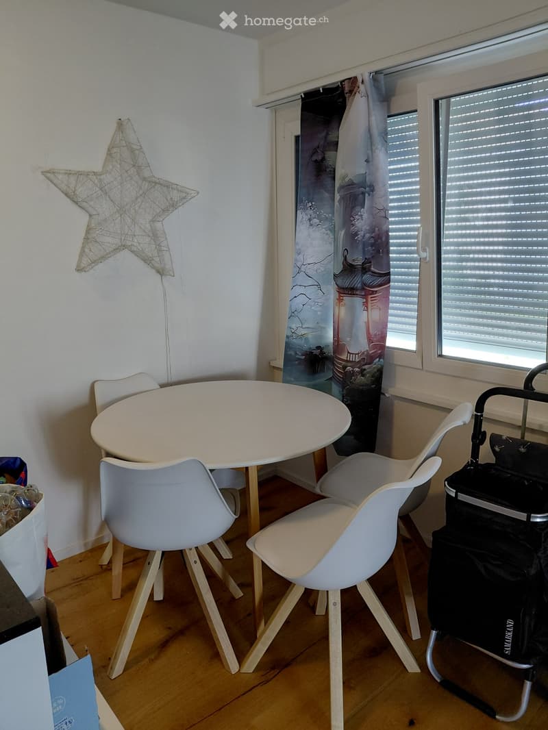 A small, well-lit room with a round white table and four white chairs, a decorative star-shaped wall hanging, and a window with blinds and a colorful curtain.