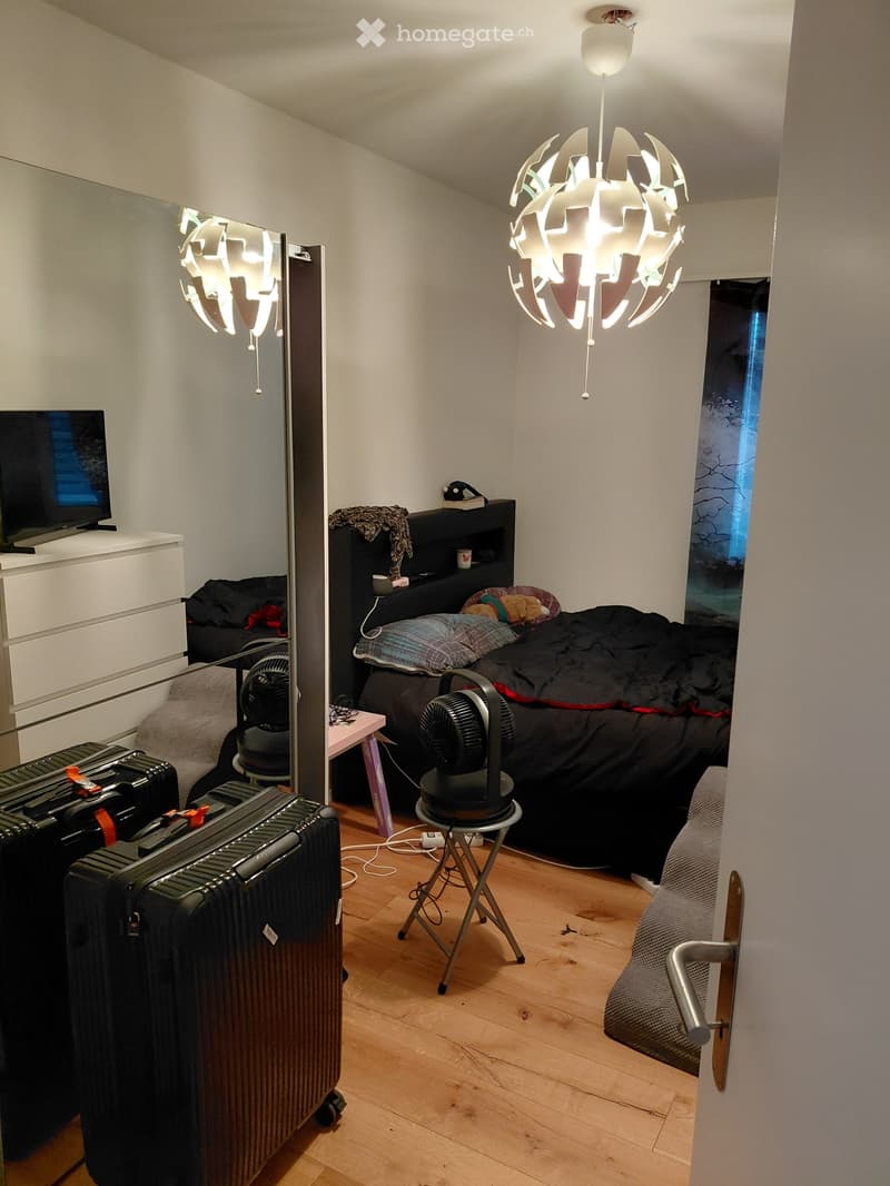 Cozy bedroom with a bed, fan, mirror, table, chair, suitcase, and a lamp. The room features wooden flooring and a modern design.