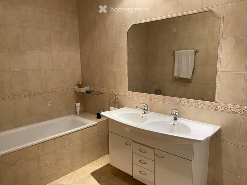 Bathroom with beige tiles, a bathtub, a double sink with a cabinet, a large mirror, and a towel hanging on a rack.
