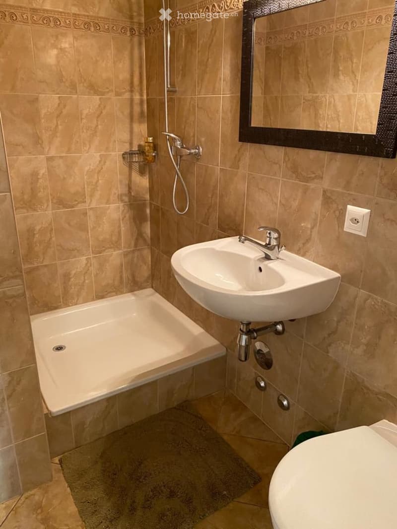 Modern bathroom with beige tiles, a bathtub, a sink with a modern faucet, a mirror, a toilet, and a shower hose with a handheld showerhead. A towel rack is mounted on the wall.
