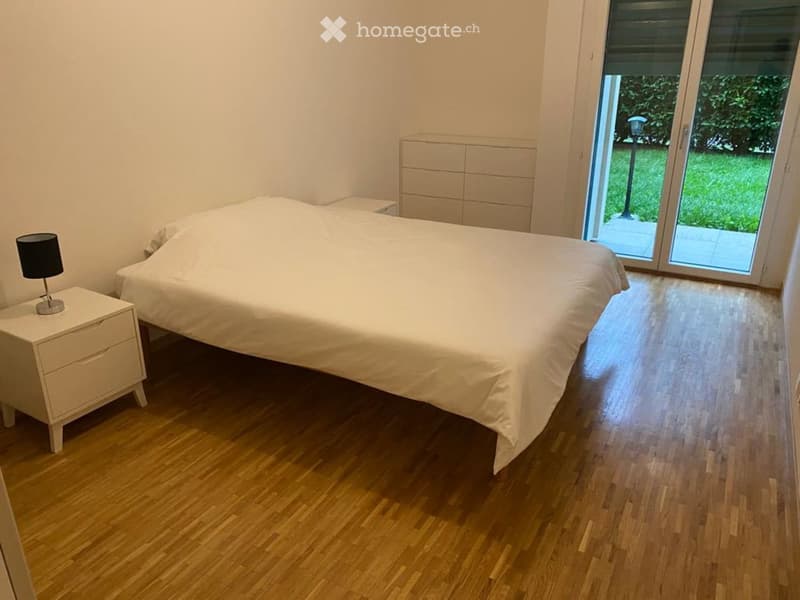 A modern bedroom with a large bed, white bedding, a nightstand with a lamp, and a headboard. The room features wooden flooring and large windows with a view of a green outdoor area.