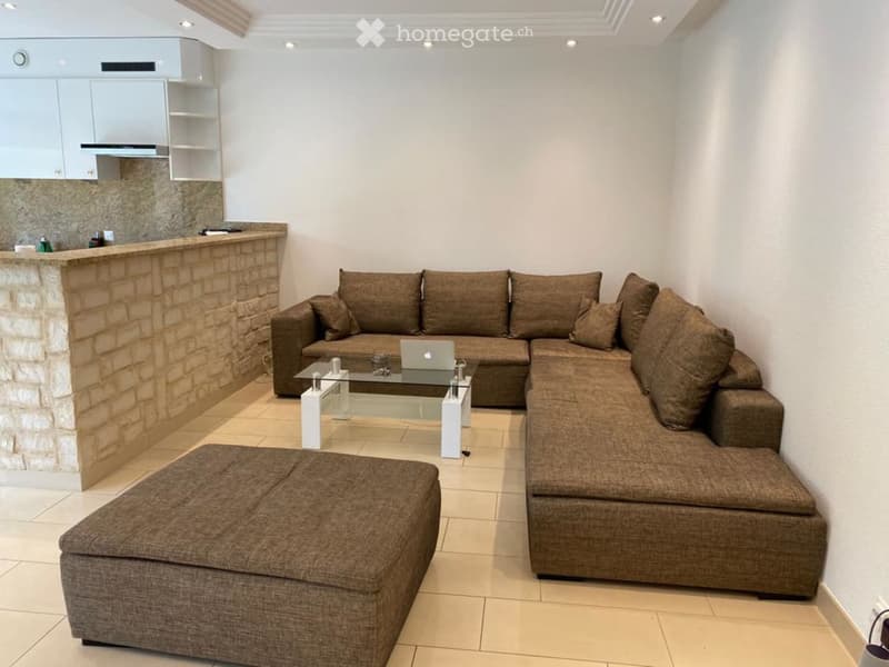 Modern living room with a brown sectional sofa, glass coffee table, ottoman, and a small kitchen area with a bar counter.