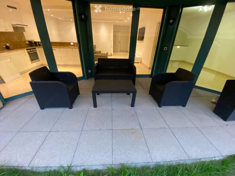 Balcony with tiled floor, black wicker furniture including sofa, table, and chairs, with a view into the living room and kitchen area.