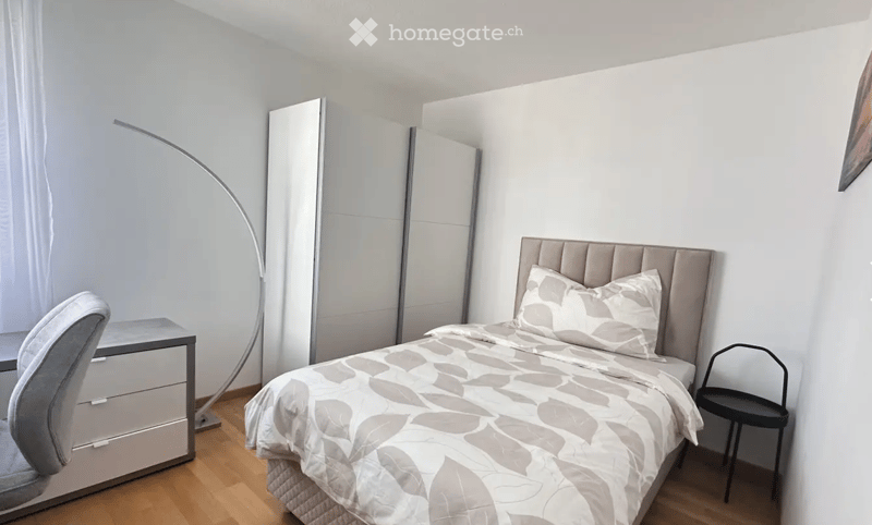 Modern bedroom with a large bed, grey and white bedding, a sleek wardrobe, a desk with a chair, and a floor lamp.
