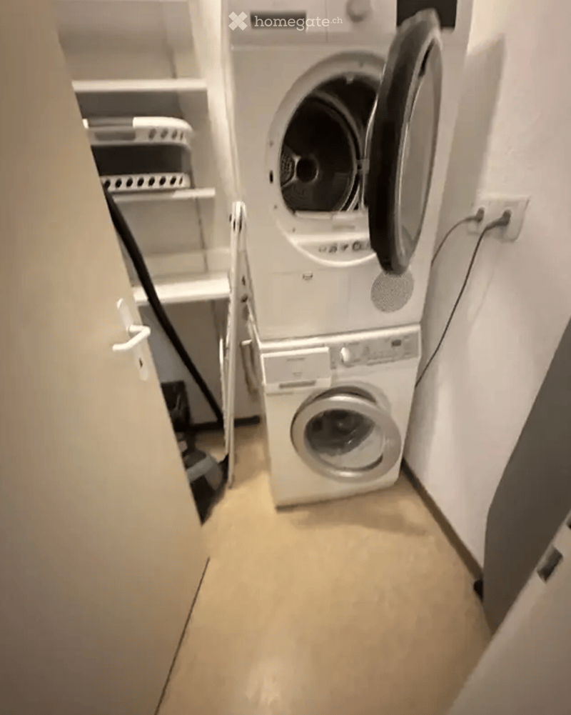 A compact bathroom with a stacked washing machine and dryer unit, white tiled walls, and built-in shelves for storage.
