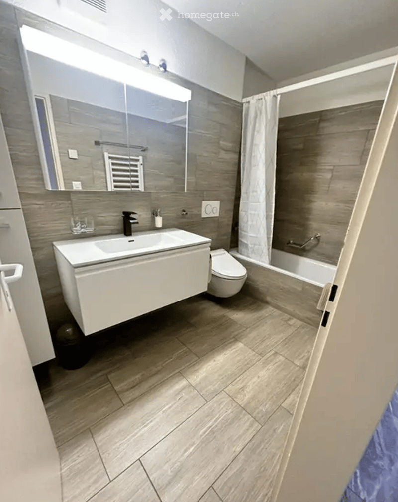Modern bathroom with a white sink, large mirror, toilet, and bathtub with a curtain. The floor is tiled in a light wood pattern, and the walls are covered in grey tiles.