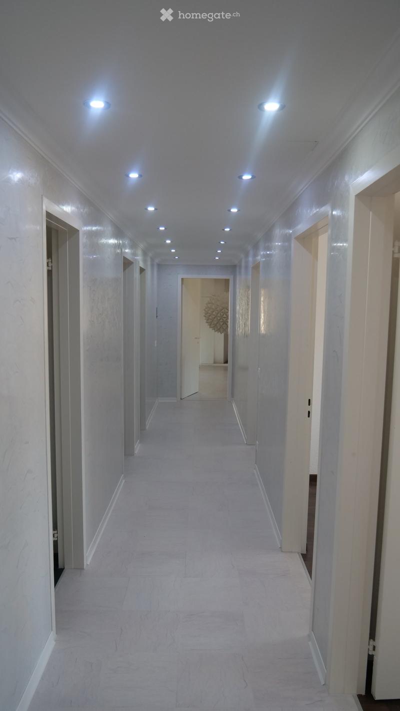 Clean, modern hallway with white marble walls and floor, recessed ceiling lights, and several open doors leading to other rooms.