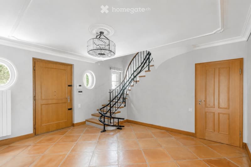 Elegant hallway with a curved staircase, wooden doors, and a chandelier. The space features light grey walls, terracotta tiles, and a decorative ceiling.