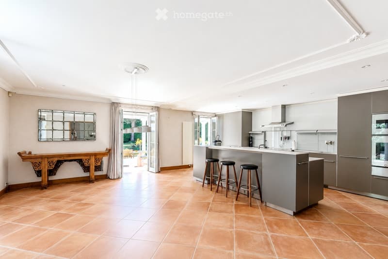 Modern kitchen with a central island, four stools, a large mirror, a wooden table, and a view to the garden through large windows.