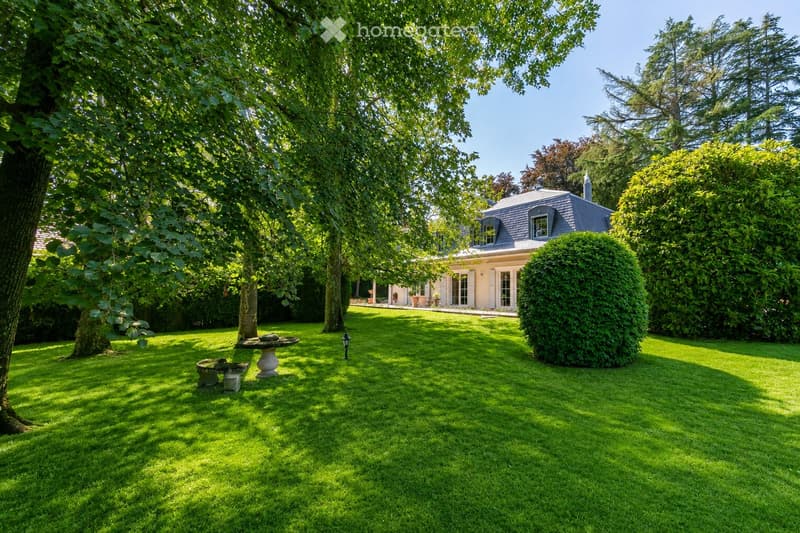 Well-maintained garden with a lawn, trees, a stone bench, and a decorative fountain. A house with a gray roof and white walls is visible in the background.