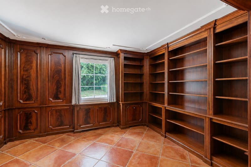 An empty room with wooden built-in bookshelves, a window with curtains, and a tiled floor.