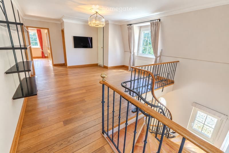 Spacious hallway with wooden flooring, a staircase with a wooden handrail and metal balustrade, a wall-mounted TV, and a chandelier light fixture.