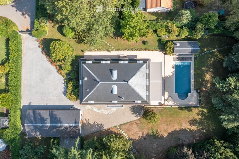 Aerial view of a two-story house with a modern roof design, surrounded by a well-maintained garden, driveway, and a swimming pool.