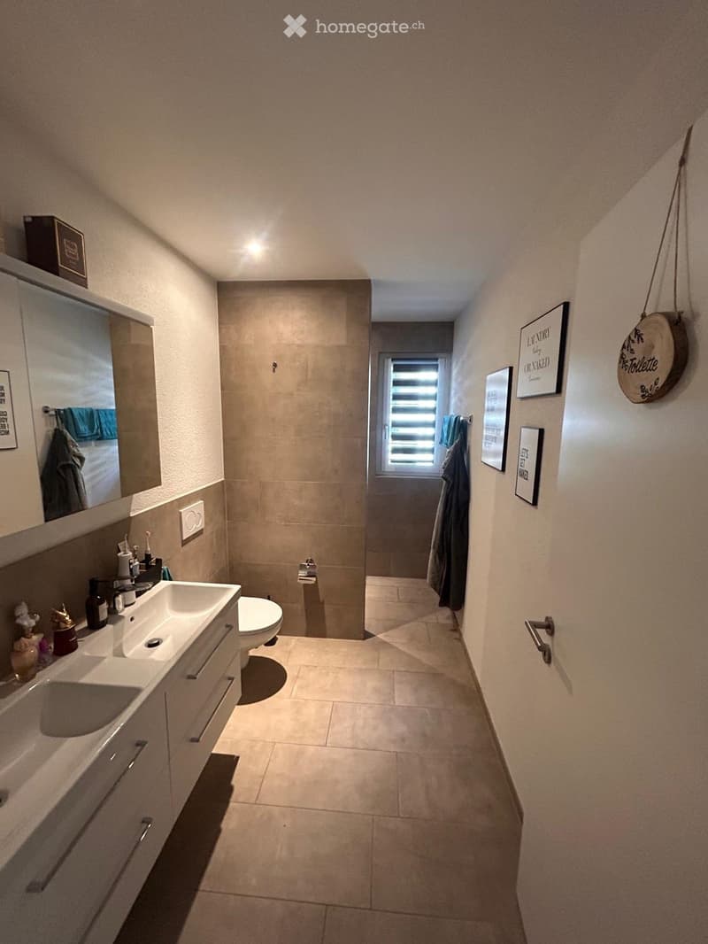 Modern bathroom with a double sink, toilet, mirror, and framed pictures on the walls. The space features beige tiles and a window with blinds.