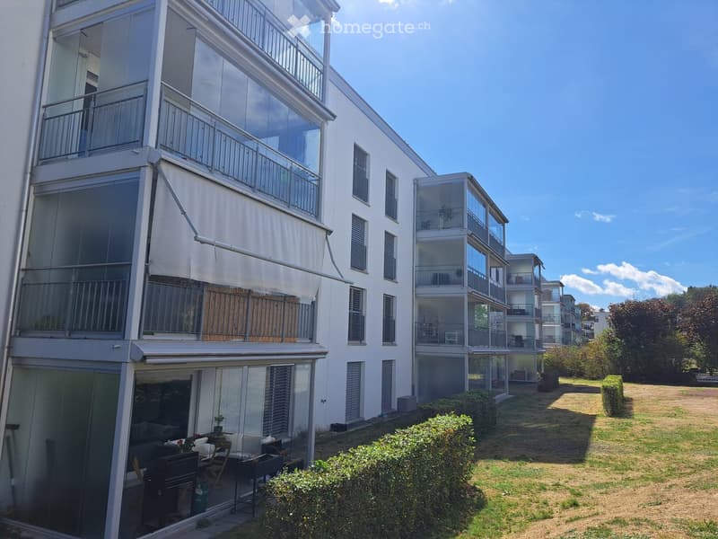 Modern multi-story residential building with large glass balconies and white exterior, surrounded by greenery and a small lawn area.