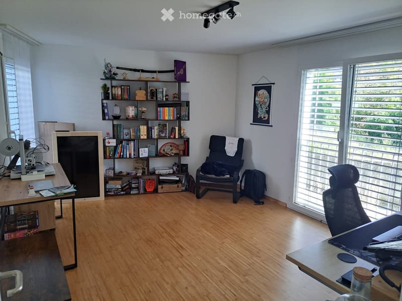A well-lit study or home office with wooden flooring, large windows, a bookshelf filled with books and decorative items, a desk with a laptop, and a comfortable chair.