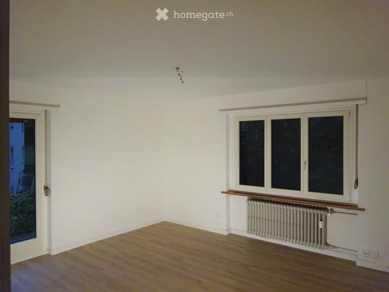 Empty room with wooden flooring, white walls, and large windows. Features a radiator and a small wooden shelf under one window.