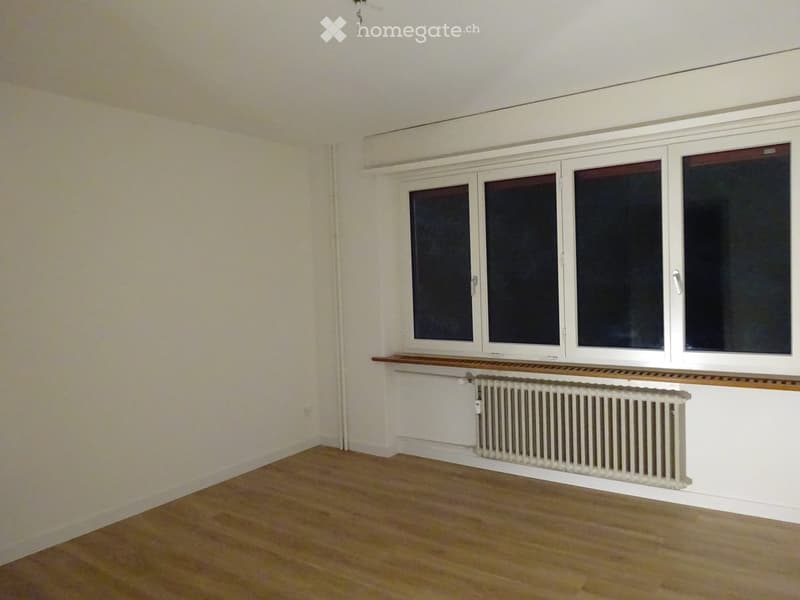 Empty room with wooden flooring, white walls, large windows, and a radiator below the windows.