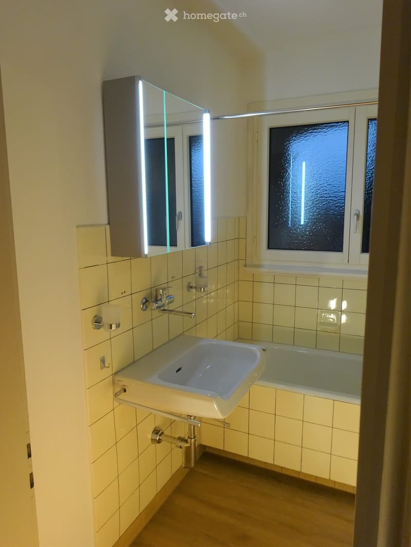 Modern bathroom with a white sink, chrome faucet, mirrored cabinet with LED lighting, and a bathtub. The walls are tiled in light yellow, and the floor is wooden.