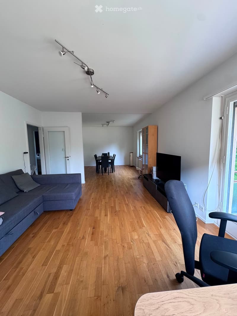 Open-plan living and dining area with wooden flooring, a grey sectional sofa, a dining table with chairs, a TV on a stand, and a wooden cabinet.