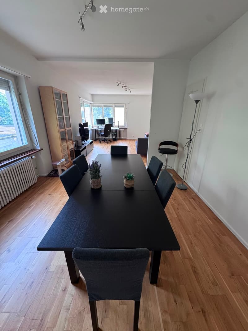 Dining room with a long black dining table surrounded by grey chairs, a floor lamp, a cabinet, and a small workspace area with a desk and computer.