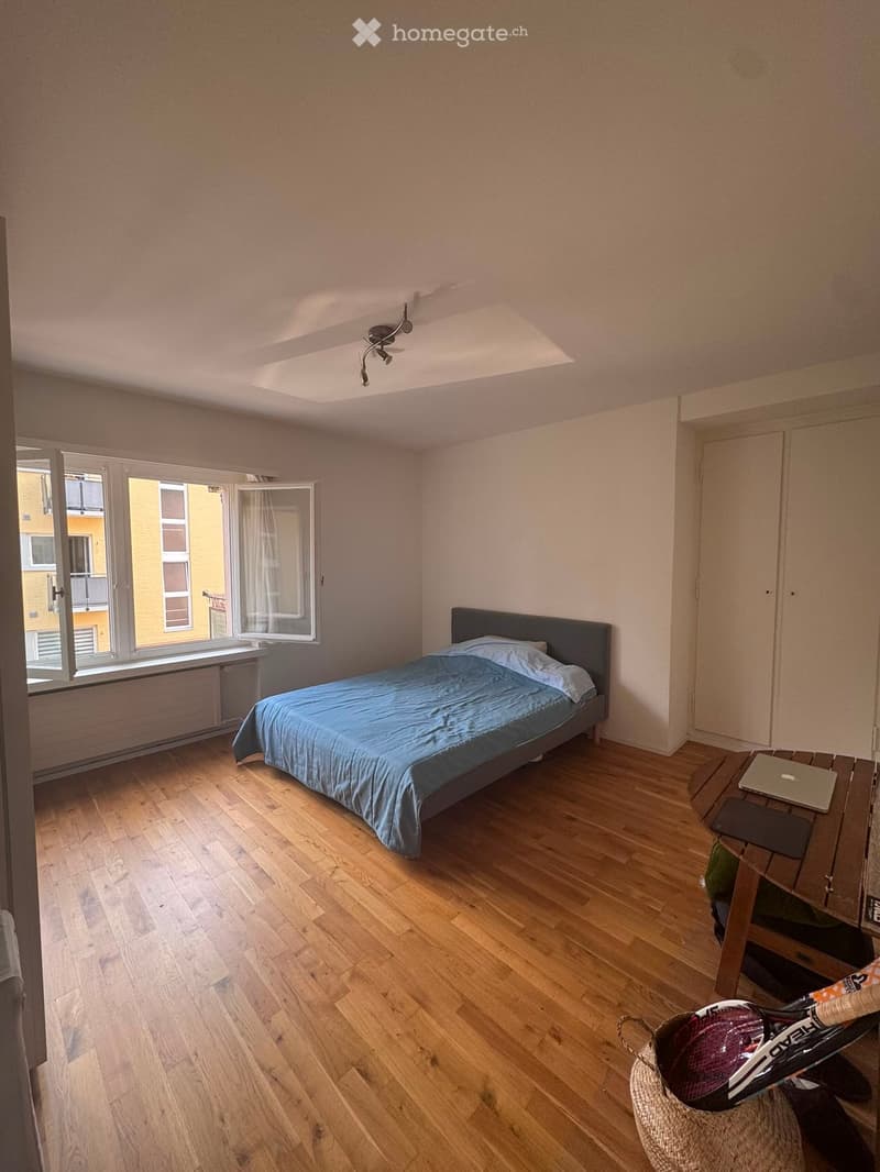 Cozy bedroom with wooden flooring, a single bed with blue bedding, a small table with a laptop, and a woven basket on the floor.
