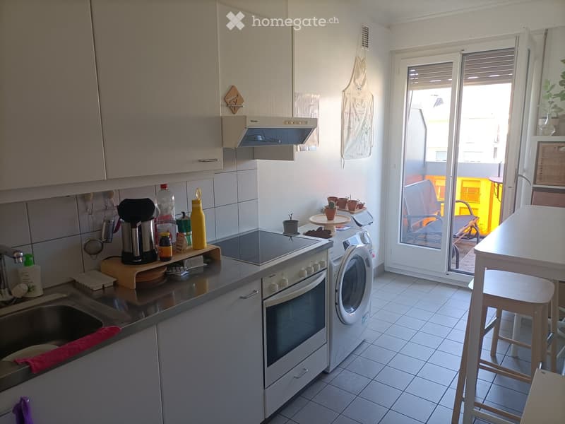 Modern kitchen with white cabinets, stainless steel appliances, and a small dining area with a table and stools. A sliding glass door leads to a balcony with outdoor furniture.