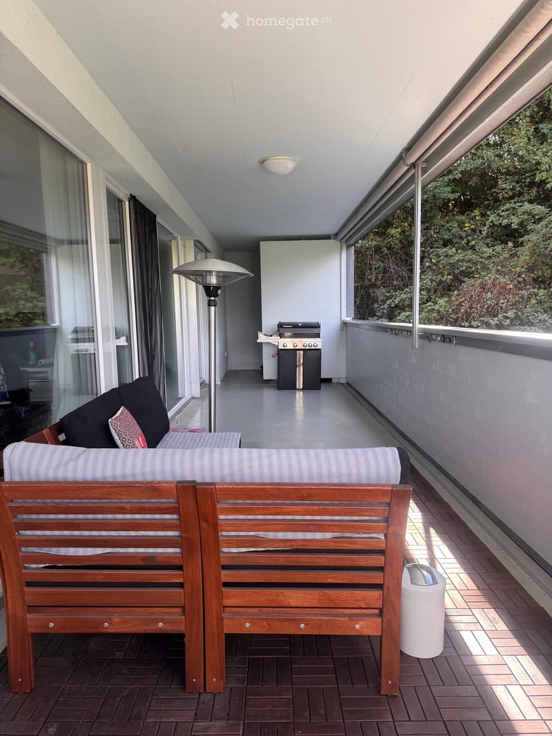 Balcony with wooden bench, sofa, grill, floor lamp, and trash can, overlooking greenery.