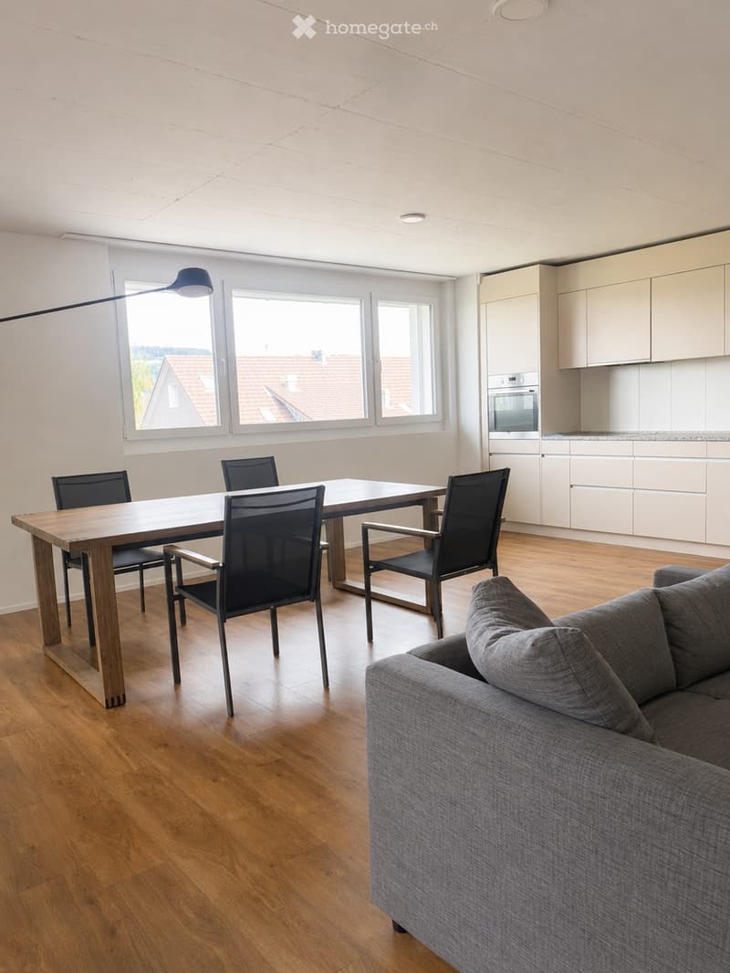 Open-plan living and dining area with a grey sofa, wooden dining table, four chairs, and a modern kitchen with white cabinets and an oven.