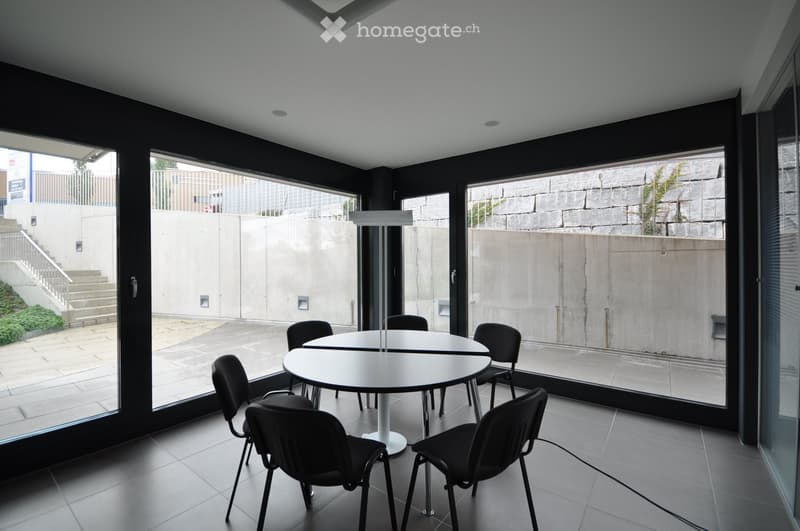 Modern, minimalist room with large glass windows, white table, and black chairs arranged around it.