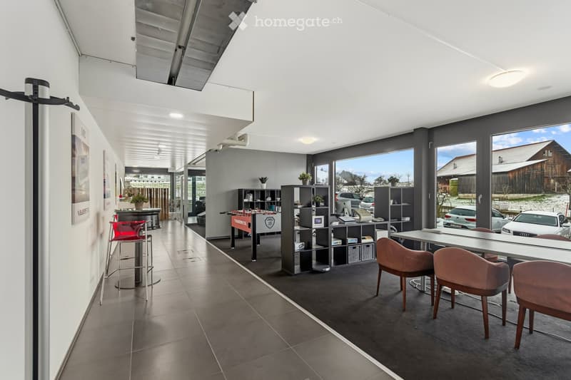 Modern office space with large windows, grey carpet, white walls, and contemporary furniture including a reception area, meeting table, and storage shelves.