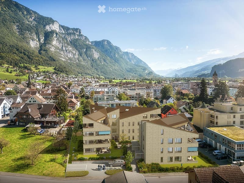 A modern multi-story building with a contemporary design, situated in a picturesque town surrounded by lush green hills and mountains.