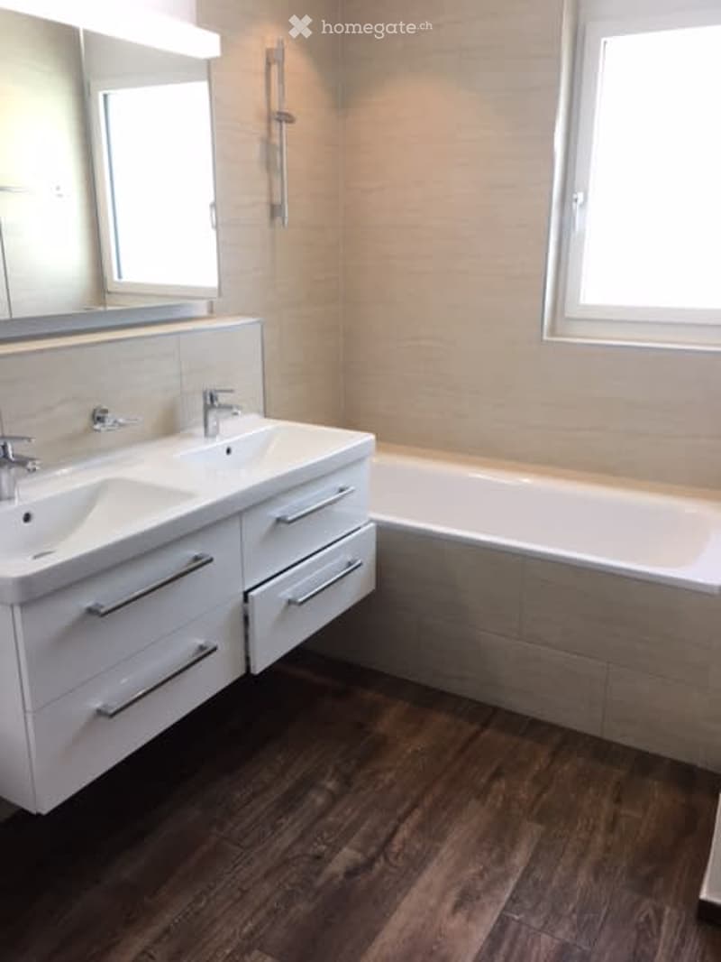 Modern bathroom with double sink vanity, bathtub, large mirror, and wooden flooring.