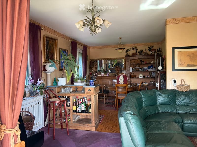 A well-furnished living room with a green leather sofa, wooden bar counter, dining table, chairs, decorative items, and artwork.