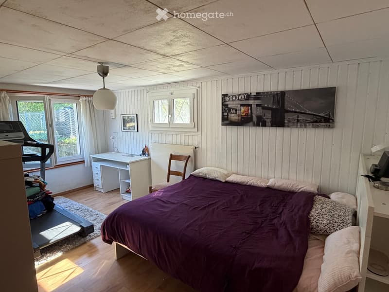 Cozy bedroom with a wooden bed, purple bedding, a desk, chair, and a large window letting in natural light. A large poster of a cityscape adorns the wall.