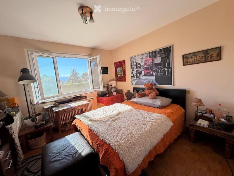 Cozy bedroom with a large window offering a scenic view, a neatly made bed with a fur throw, nightstands with lamps, a framed picture, and a small table with a plant.