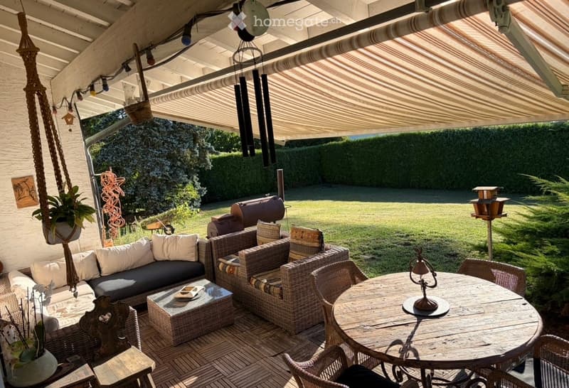 A covered outdoor patio with a sofa, wicker chairs, a wooden table, and hanging decorations. The space features a wooden deck, a barbecue, and a view of a grassy lawn.