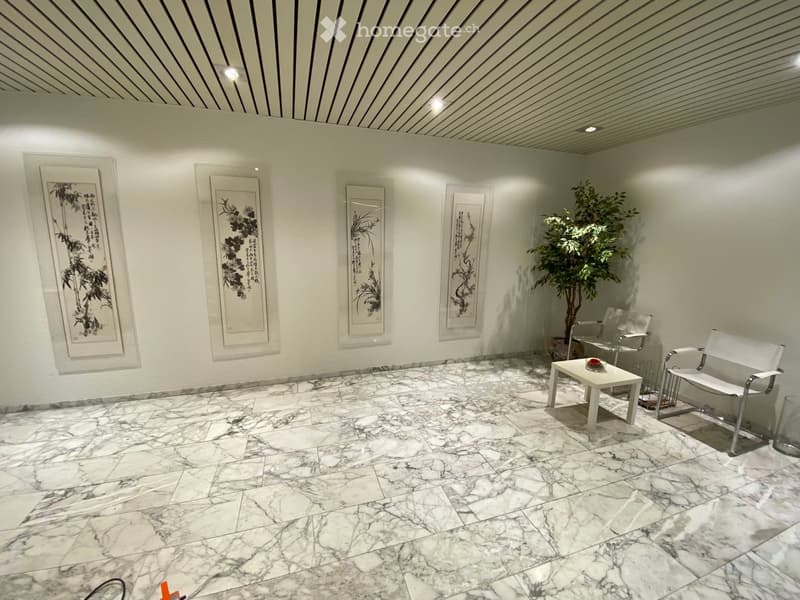 A modern, minimalist room with marble flooring, white walls, and framed artwork. Contains a small white table, two white chairs, and a potted plant.