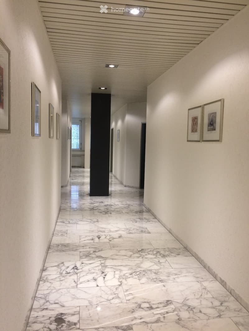 A long, well-lit hallway with marble flooring, white walls, and framed artwork on both sides. The ceiling features recessed lighting and a decorative panel design.