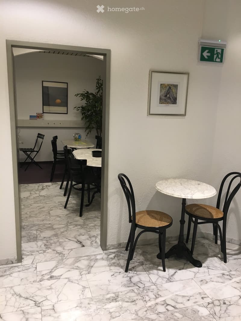 A small, furnished area with a round marble table, two black chairs, and a potted plant. An adjacent room features additional tables and chairs.